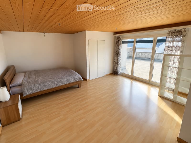 Bright bedroom with wooden flooring, a single bed with pillows and a blanket, a nightstand with a vase, a white shelf, and a wardrobe. Large windows with curtains lead to a balcony.