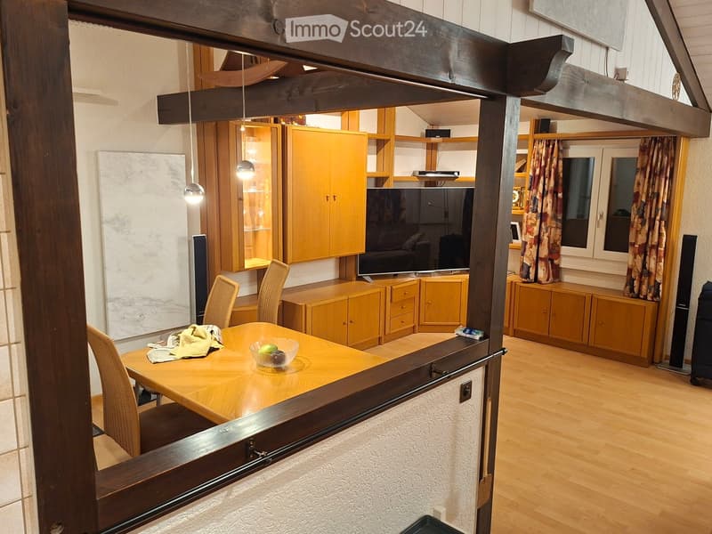 A well-lit living and dining area with wooden flooring, a dining table with chairs, cabinets, a TV screen, and curtains on the windows.
