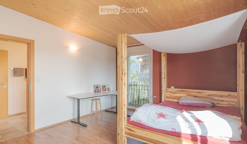 Cozy bedroom with wooden bed, red bedding, a small table with a stool, and a window with a view of the garden.
