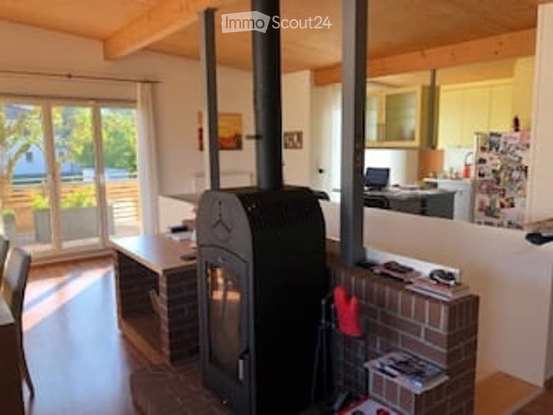 The image shows a cozy living room with a wooden ceiling and floor, a wooden table and chairs, a wood-burning fireplace, and a kitchen area with a refrigerator and a laptop on the counter.