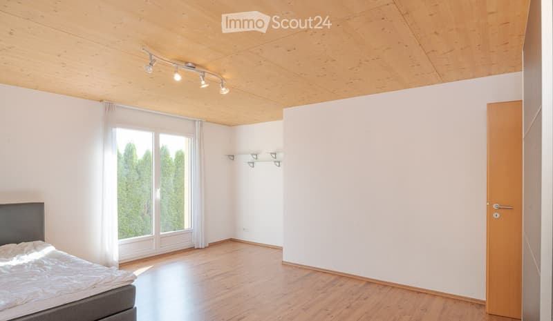 Bright bedroom with wooden floor, white walls, large window with curtains, and built-in shelves. The room features a modern bed and a sleek wooden door.