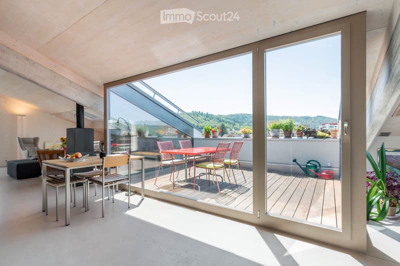 Bright living room with large glass doors opening to a balcony, featuring a sofa, chair, small table, and fireplace. The balcony has a dining table with chairs and potted plants.