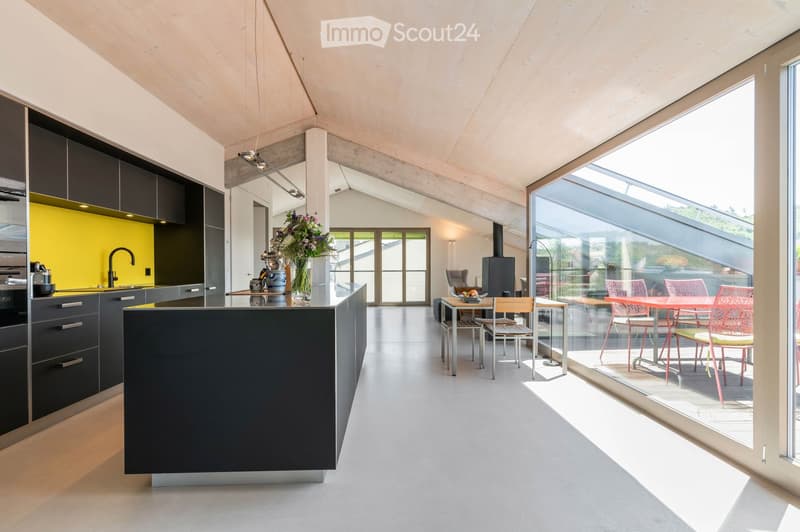 Modern kitchen with dark grey cabinets, a central island, and a dining area with a table and chairs. The space features large windows and a sleek design.