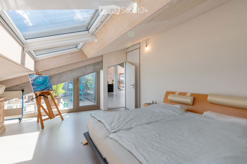Bright, modern bedroom with a large bed, wooden headboard, skylight windows, and an open doorway leading to a balcony area with outdoor furniture.