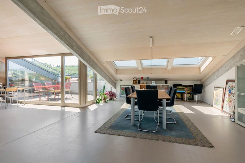 Bright, open dining and living area with large windows and skylights, featuring a wooden dining table with black chairs, a sofa, a small table with chairs, and a potted plant near the window.