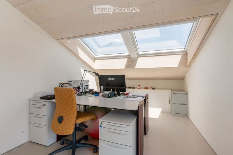 A modern, well-lit attic office space with a slanted wooden ceiling and skylights. The room features a white desk with a computer monitor, a comfortable orange office chair, a printer, and various office supplies. There is also a small cabinet and a red e