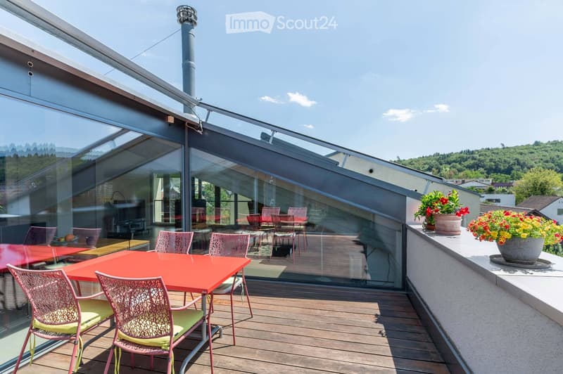 Modern rooftop terrace with wooden decking, red table, pink chairs, and potted flowers, offering scenic views of the surrounding landscape.