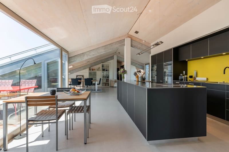 Modern kitchen with a central island, dark cabinets, and a bright yellow accent wall. The space includes a dining area with a wooden table and chairs, and large windows allowing natural light to fill the room.