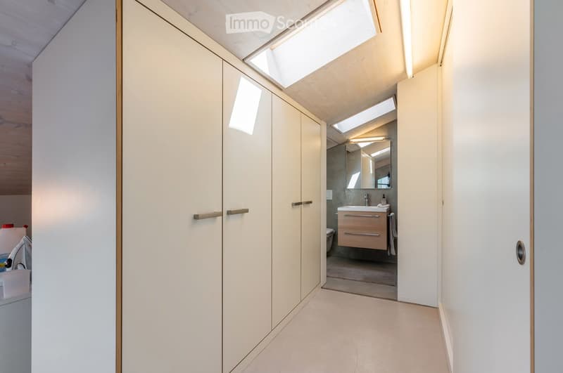 A modern hallway with white walls, light wooden flooring, and built-in white storage units with brass handles. The hallway leads to a bathroom with a vanity, sink, and mirror.