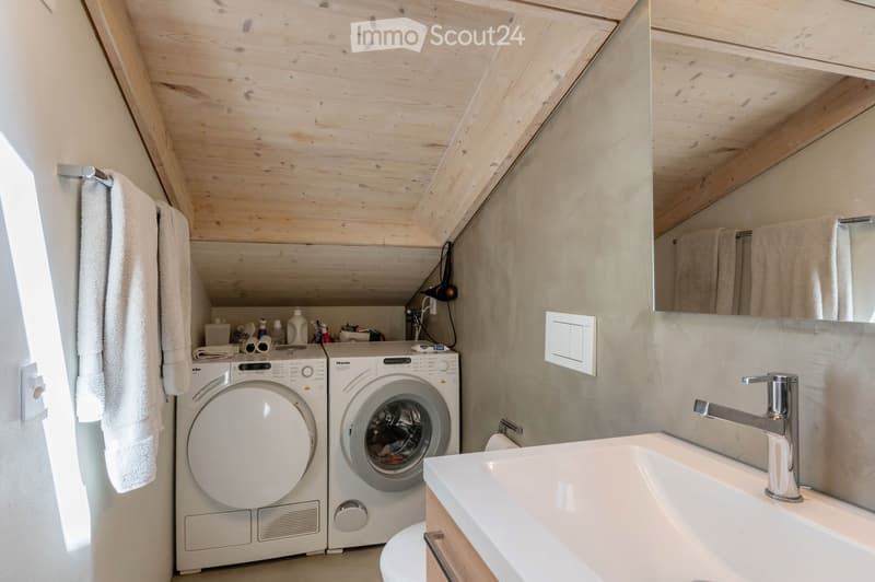 Bathroom with a sink, faucet, mirror, washing machine, dryer, and towels hanging on a rack. The room features wooden ceilings and light-colored walls.