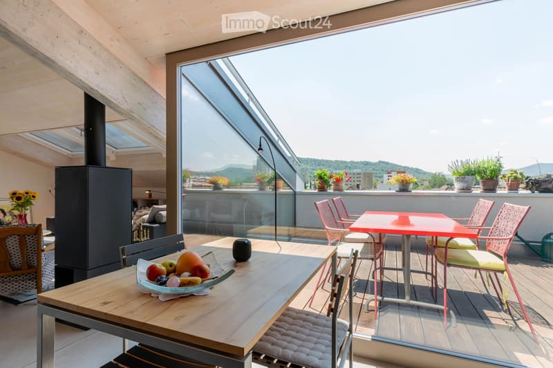 Modern rooftop terrace with large glass walls offering a scenic view, featuring a wooden dining table, colorful chairs, and potted plants.