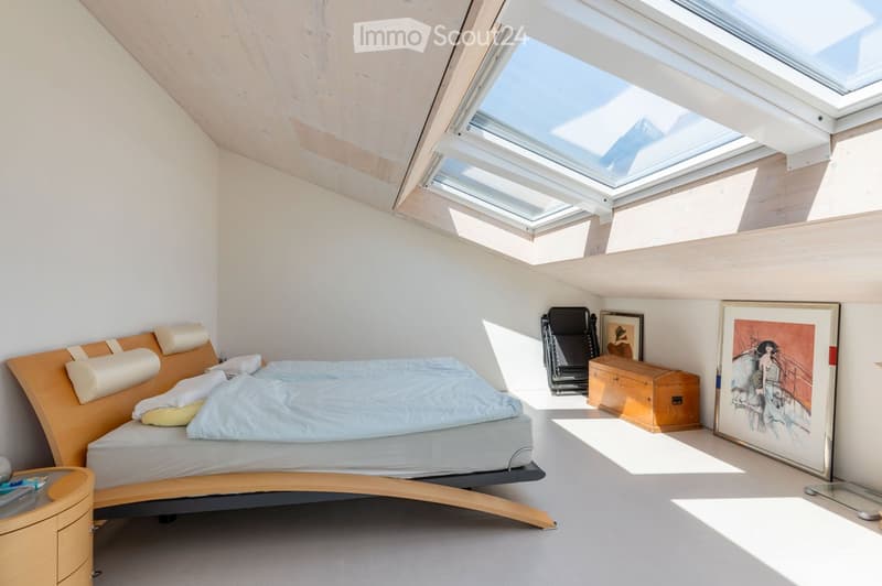 Modern bedroom with a slanted wooden ceiling and skylights, featuring a contemporary bed, nightstand, chair, chest, and artwork.