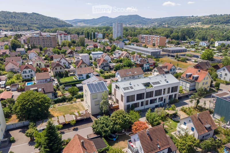 Aerial view of a suburban neighborhood with single-family homes, apartment buildings, and green spaces, surrounded by hills and trees.