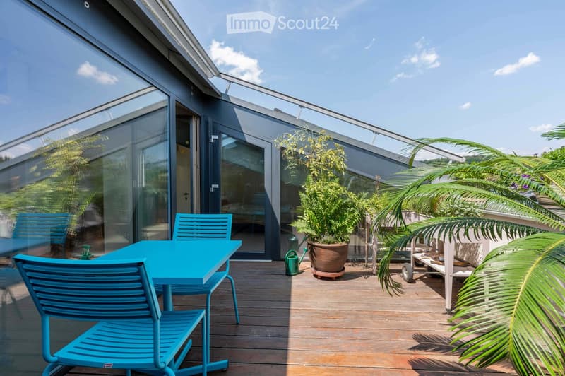 Modern rooftop terrace with wooden decking, blue table and chairs, potted plants, and a view of the building.