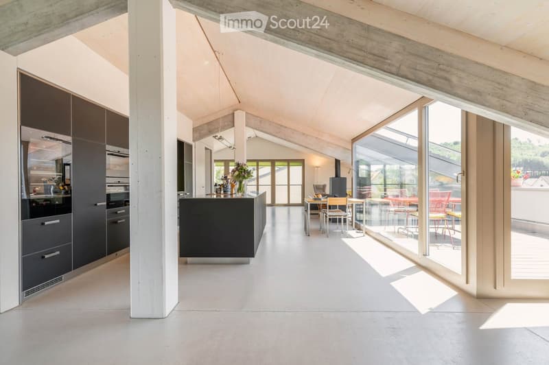 Modern kitchen with grey cabinetry, a central island, and a dining area with chairs and a table. Large windows and a concrete ceiling create an open, airy space.