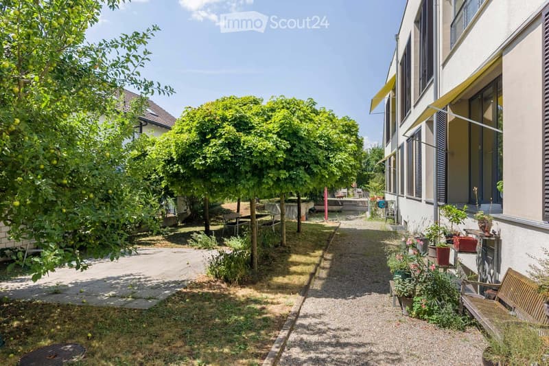 A narrow garden path lined with trees and potted plants leads to a building entrance. The area includes a wooden bench, small tables, and various greenery.