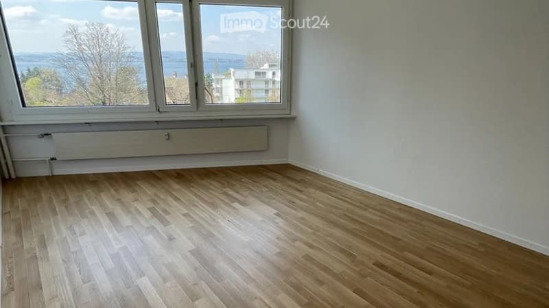 Empty room with light wooden flooring and large windows offering a scenic view of the water and surrounding area.