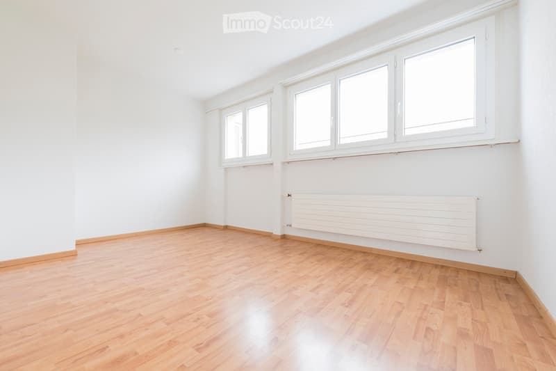 Empty room with light wooden flooring, white walls, and large windows with white frames. A white radiator is mounted on one wall.
