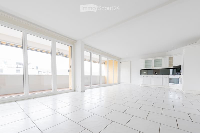 Bright, unfurnished room with white tiled flooring, large glass sliding doors leading to a balcony, and a white kitchen area with cabinets and appliances.