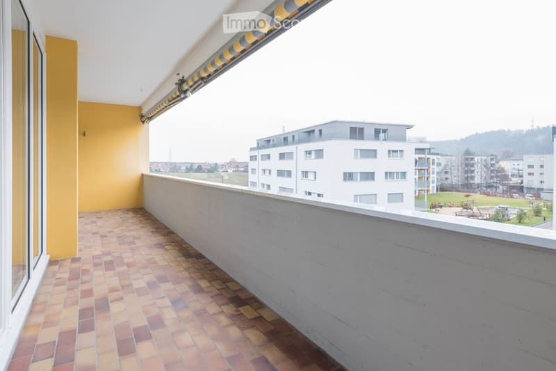 Balcony with tiled floor, yellow wall, and retractable awning, overlooking a residential area with buildings and a park.