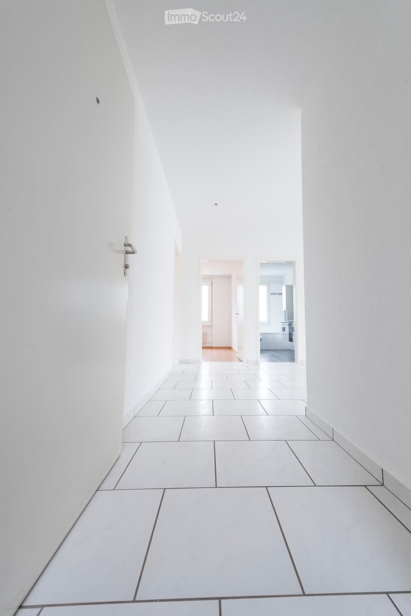 A clean, minimalist hallway with white tiled flooring and white walls, featuring a door on the left and two open doorways leading to other rooms.