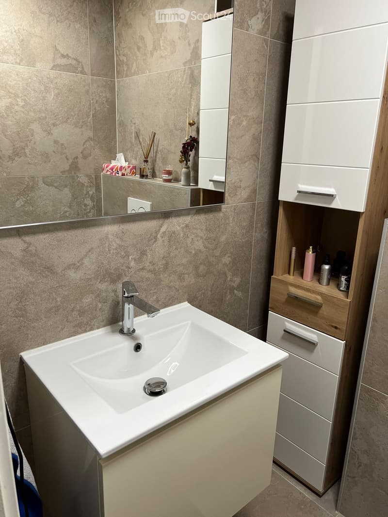 Modern bathroom with a white sink, chrome faucet, mirrored cabinet, and wooden storage unit. The countertop holds decorative items including candles, a vase with flowers, and a tissue box.