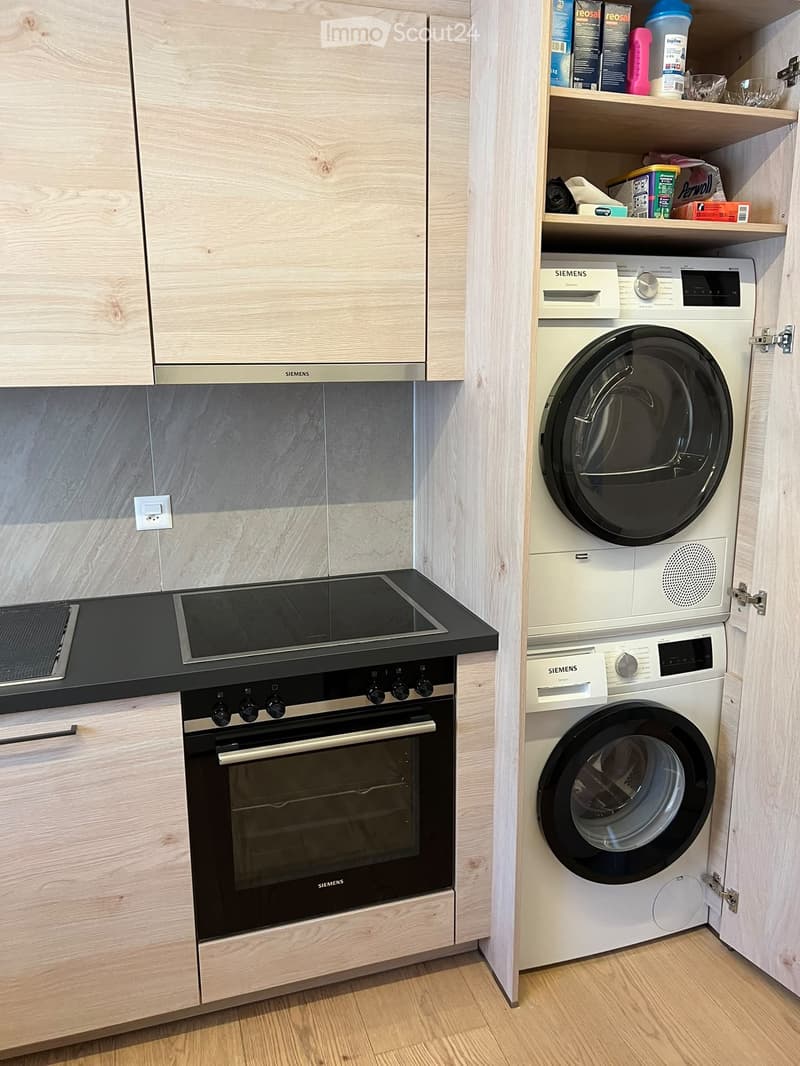 Modern kitchen with light wooden cabinets, stainless steel appliances including an oven, induction stove, and a stacked washer/dryer unit.