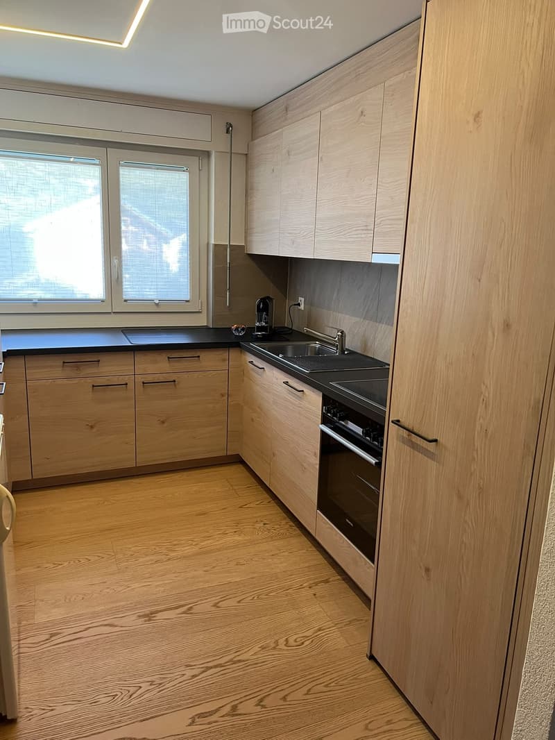 Modern kitchen with light wooden cabinets, black countertops, and a stainless steel sink and oven. Features a coffee maker on the counter and a window with blinds.