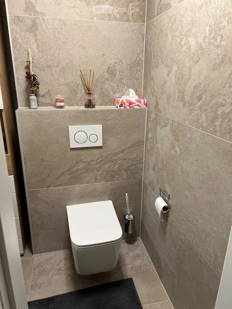Modern bathroom with beige marble tiles, white toilet, toilet paper holder, toilet brush holder, decorative vase, candles, and tissue box on the shelf.