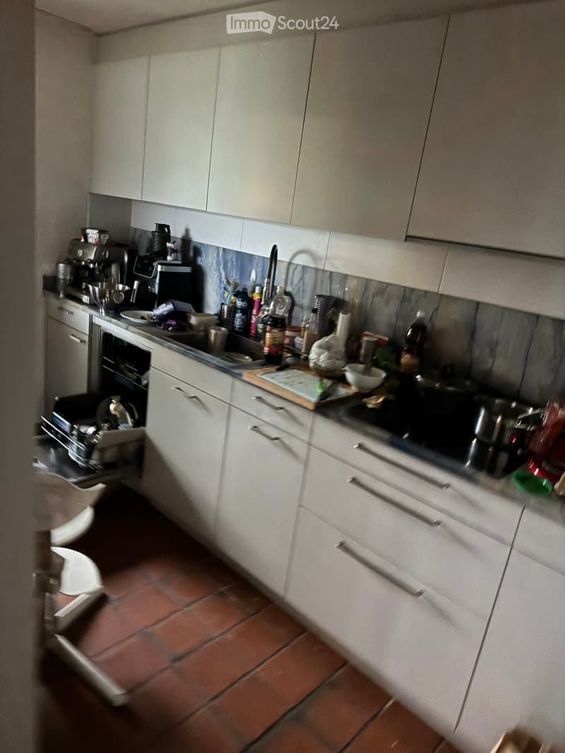 Modern kitchen with white cabinets, stainless steel appliances, and a tiled floor. The counter is cluttered with various kitchen items including a coffee maker, cutting board, frying pan, and other utensils.