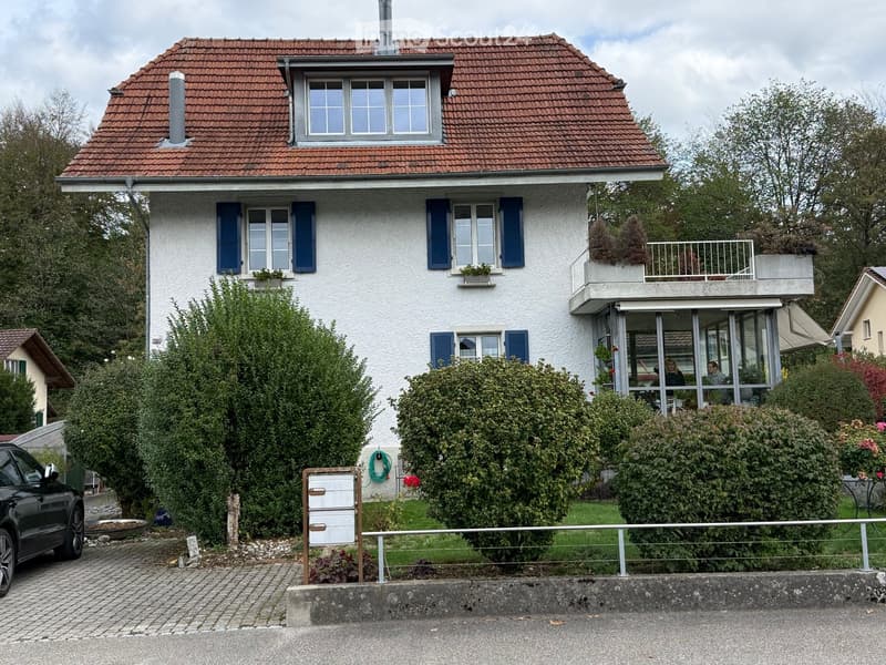 Two-story house with red-tiled roof, white exterior, blue shutters, and a balcony on the upper level. The house is surrounded by greenery and has a driveway with a parked car.