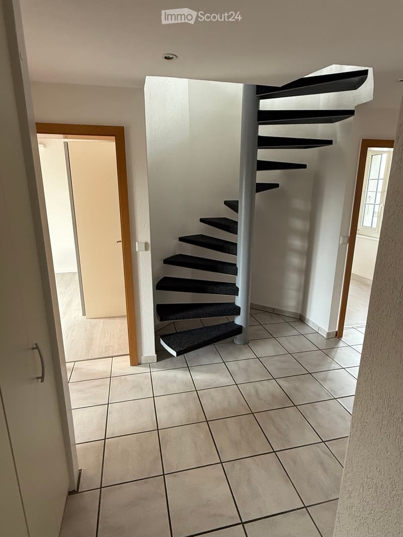 Spiral staircase with black carpeted steps and white central column, leading to an upper level. The hallway has tiled flooring and white walls, with a door visible on the left side.