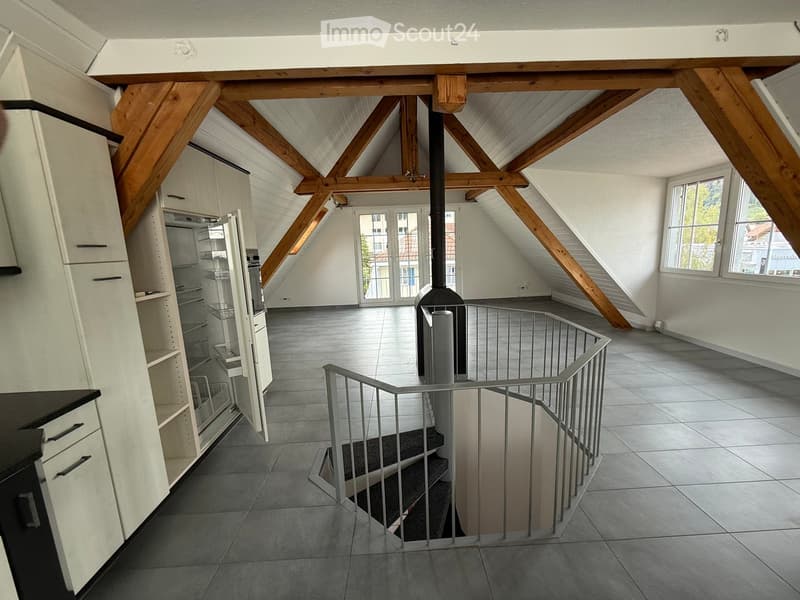 Open loft-style room with exposed wooden beams, tiled floor, metal staircase, and large windows. No furniture or appliances are present.