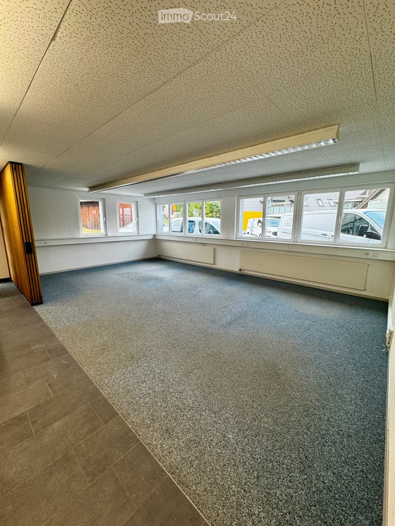 Empty room with grey carpeted floor, white walls, large windows, and a wooden door. No furniture or equipment present.