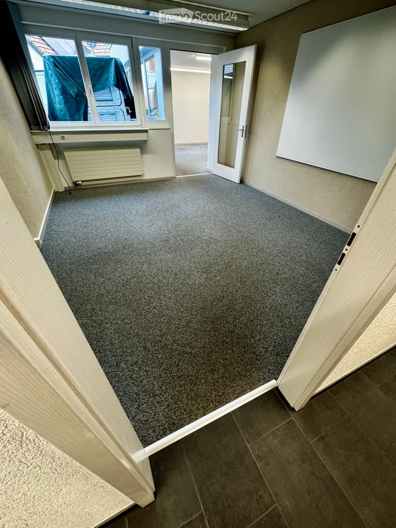 Empty room with grey carpeted floor, white walls, large windows, and an open door leading to another room.