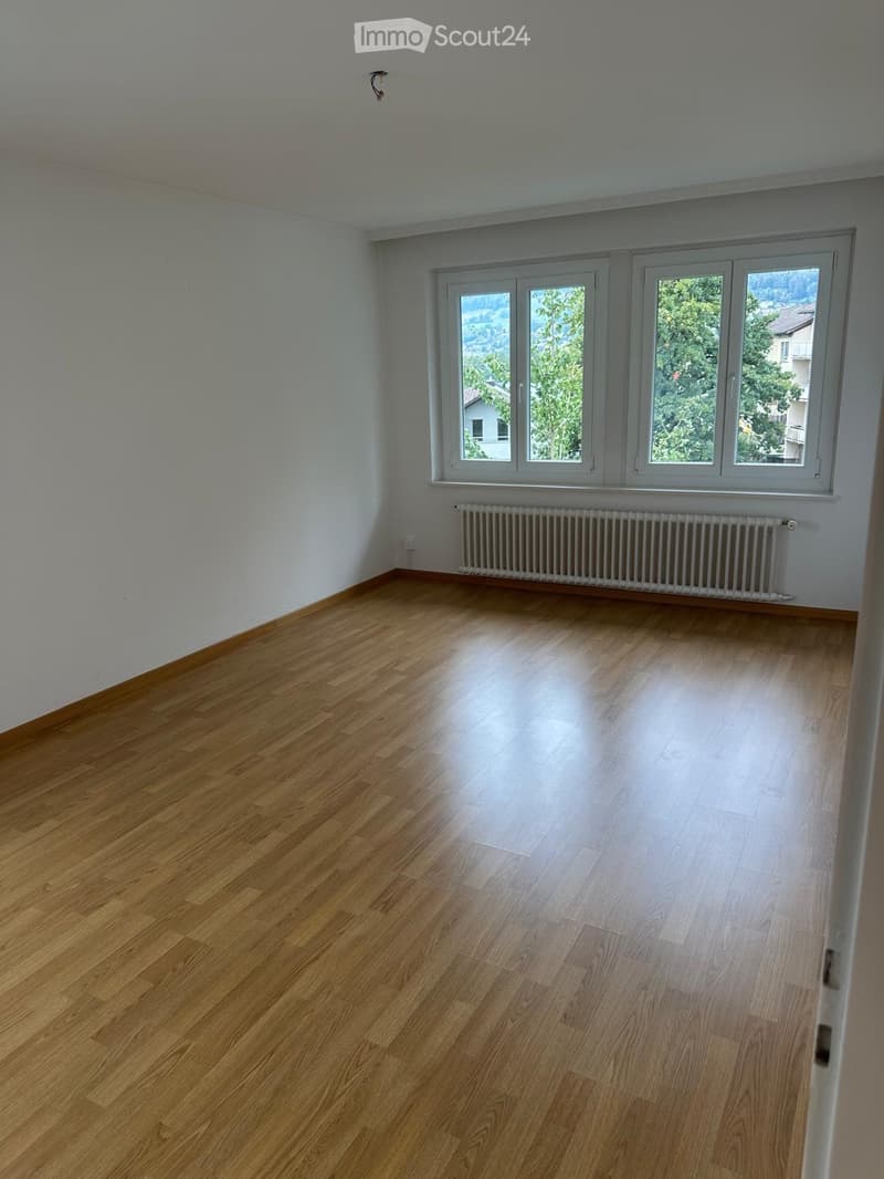 Empty room with wooden flooring, white walls, and large windows offering a view of greenery and buildings outside. A radiator is visible under the windows.