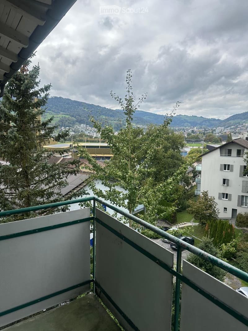 Balcony with metal railing overlooking a scenic view of a town, trees, and distant mountains.