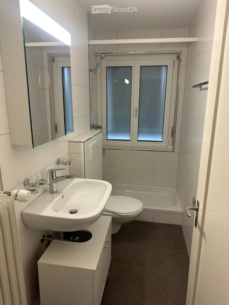 Modern bathroom with white tiles, a sink with a modern faucet, a mirror, a toilet, a bathtub, and a shower area with a glass door. The space includes a small cabinet and a window with frosted glass.