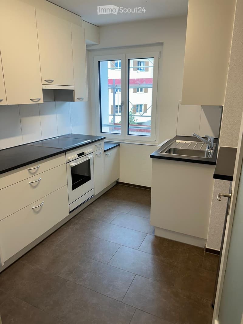 Modern kitchen with white cabinets, black countertops, a sink, and an oven. The space is well-lit with large windows offering a view of the outside.