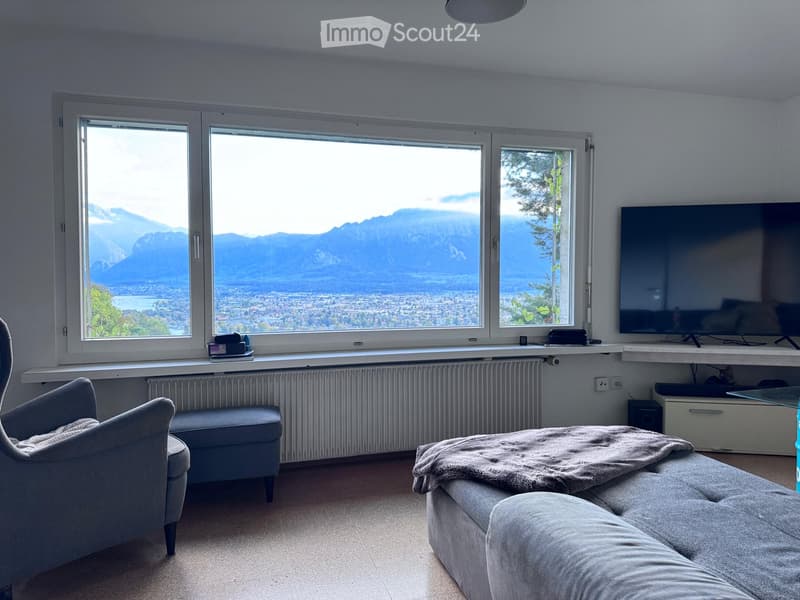 Modern living room with large windows offering a scenic mountain view, featuring a grey sofa, armchair, ottoman, and a flat-screen TV on a white shelf.