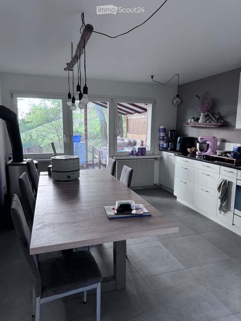 Modern kitchen with white cabinets, grey tiled floor, and a large wooden dining table with chairs. Visible appliances include an oven, coffee maker, and mixer. There is a radiator and a shelf with decorative items.