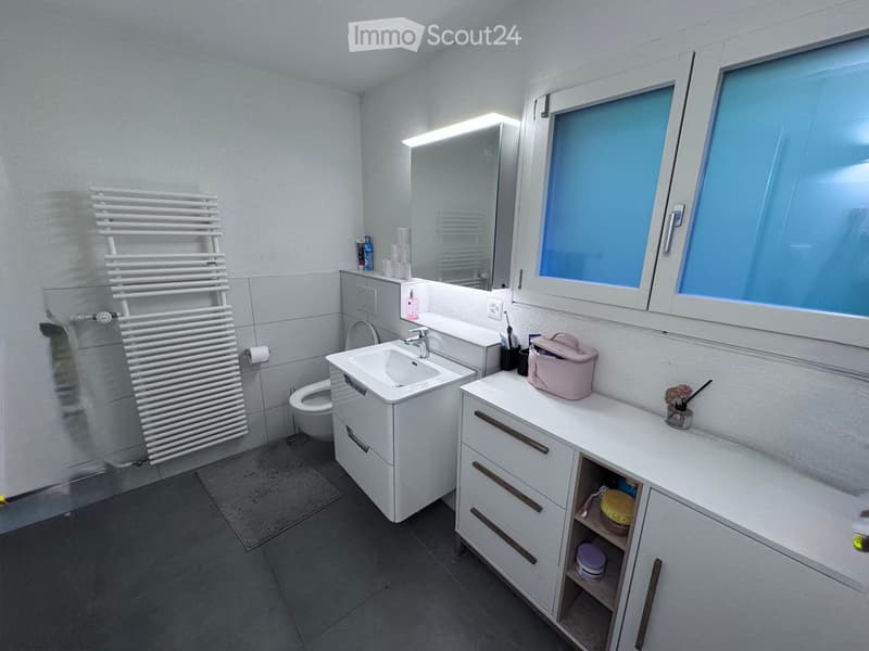 Modern bathroom with white tiles, a sink with a mirror, a toilet, a towel rack, and a small cabinet with toiletries and a flower vase on the counter.