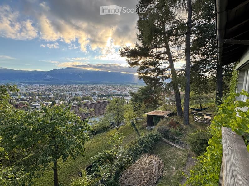 A scenic outdoor view from a garden or terrace, showing a cityscape with buildings, trees, and distant mountains under a partly cloudy sky.