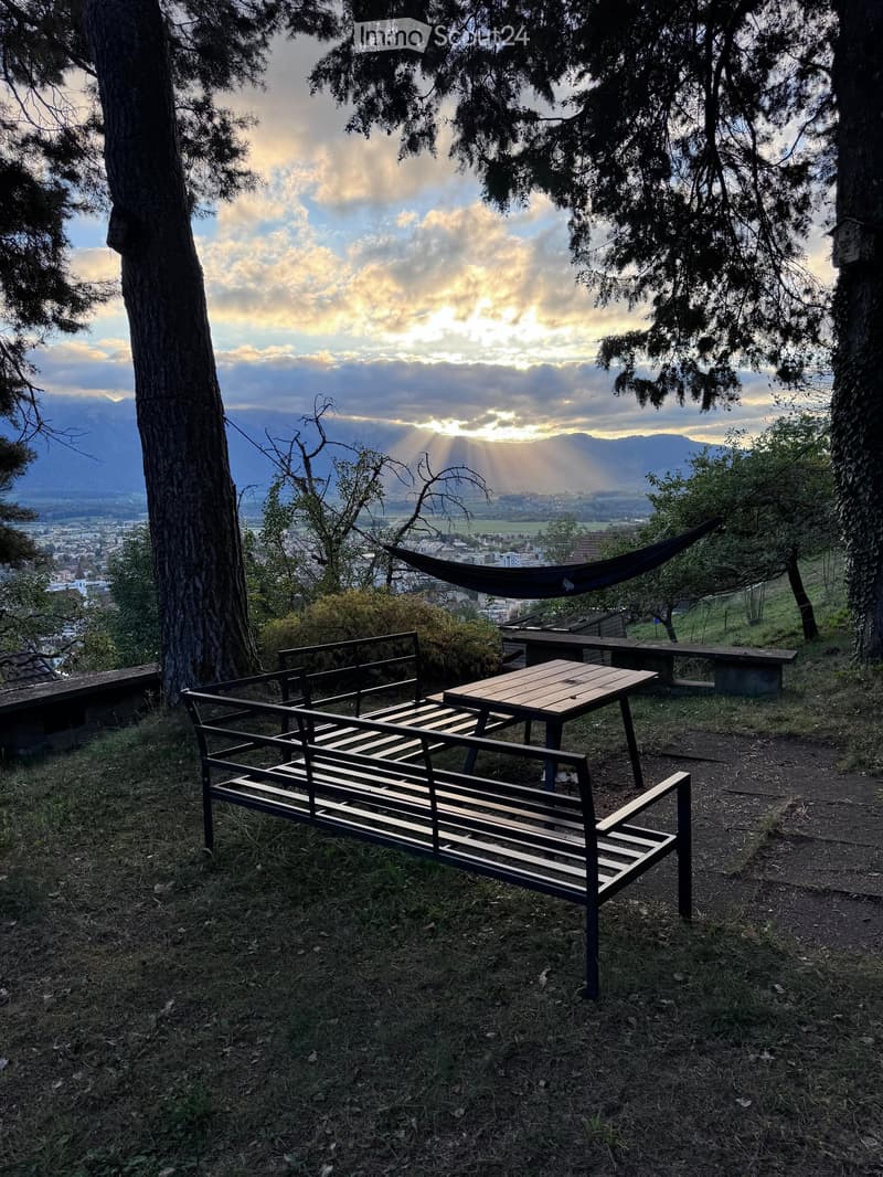 Outdoor seating area with a bench, table, and hammock, overlooking a scenic valley and mountains at sunset.