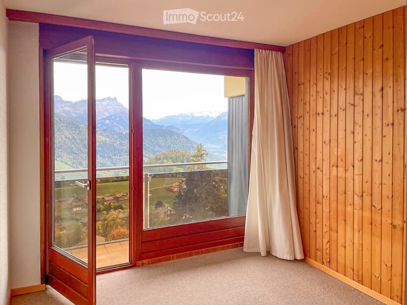 Empty room with wooden panel walls, large windows offering a scenic mountain view, and a sliding glass door leading to a balcony.
