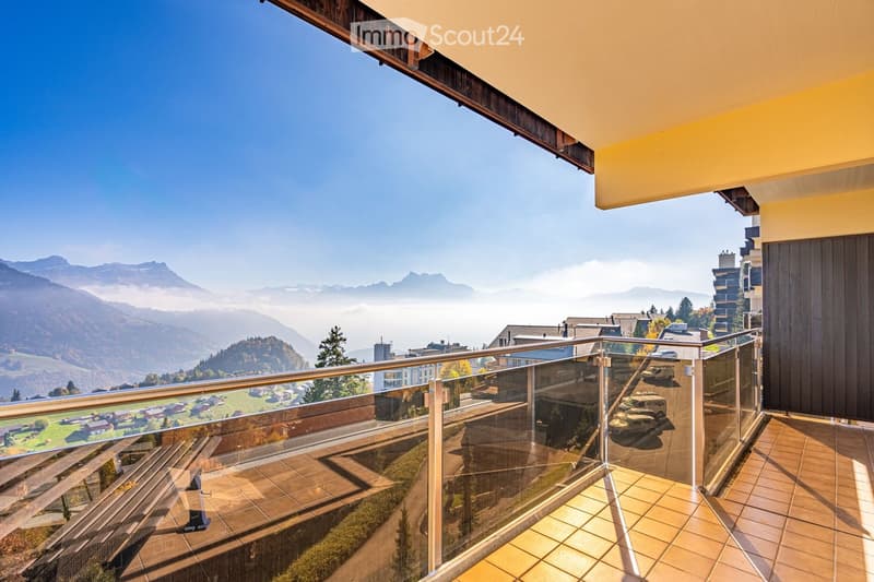Balcony with glass railing, tiled floor, overlooking a scenic valley and distant mountains.
