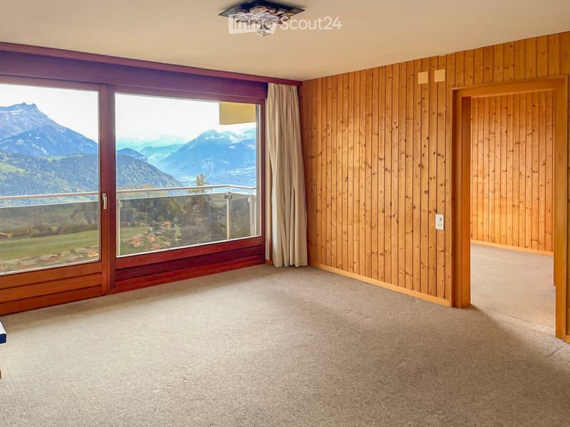 Empty room with wooden panel walls, large windows offering a scenic mountain view, and carpeted floor.