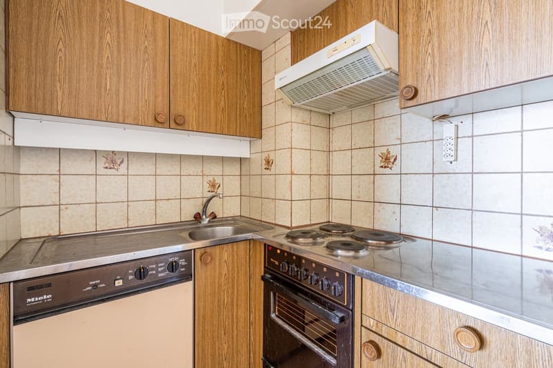 A compact kitchen with wooden cabinets, a stainless steel sink, a four-burner stove, and an oven. The walls are tiled with a floral pattern, and a range hood is mounted above the stove.