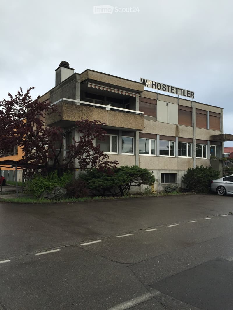 Two-story building with a balcony, signage reading "W. HOSTETTLER", surrounded by greenery and a parking area.