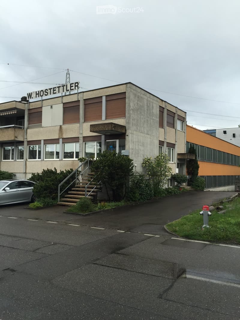 Two-story building with concrete facade, brown shutters, and a sign reading "W. HOSTETTLER" on the roof. A silver car is parked in front, and there is a small staircase leading to the entrance.